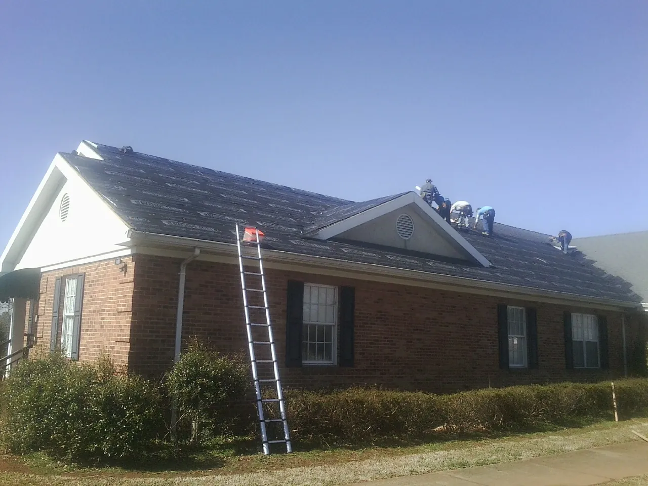 Crew tearing shingles off a brick ranch roof with a ladder set — Emergency Roof Repair work by our crew in Autaugaville
